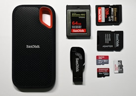 An assortment of storage devices is displayed, including a black SanDisk portable SSD with a red accent, several memory cards such as SD and microSD of varying capacities from brands like SanDisk and Transcend, along with a USB flash drive and a microSD card adapter.