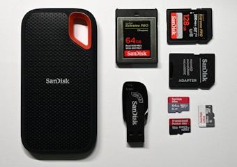 An assortment of storage devices is displayed, including a black SanDisk portable SSD with a red accent, several memory cards such as SD and microSD of varying capacities from brands like SanDisk and Transcend, along with a USB flash drive and a microSD card adapter.
