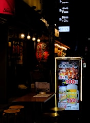 A dimly lit street scene featuring a Japanese restaurant with bright lanterns and illuminated signage. A small table with stools is visible in the foreground, while colorful signs with Japanese text advertise food and drink specials, including images of grilled chicken and beer.