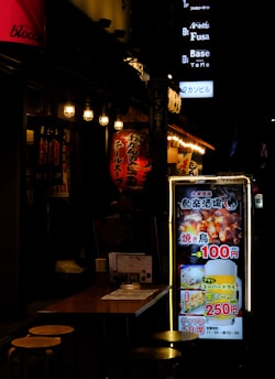 A dimly lit street scene featuring a Japanese restaurant with bright lanterns and illuminated signage. A small table with stools is visible in the foreground, while colorful signs with Japanese text advertise food and drink specials, including images of grilled chicken and beer.