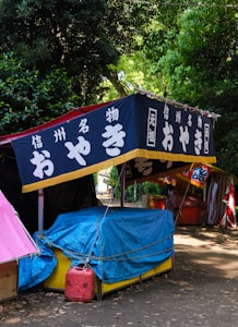 A makeshift stall is covered with a blue tarp, surrounded by lush greenery. A red gasoline can is placed on the ground, and the stall is adorned with a sign featuring Japanese text on a dark background with a yellow border.