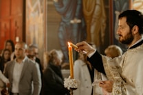 A man in ornate robes is lighting a candle in what appears to be a religious setting, with several people gathered in the background. The scene is indoors with warm lighting and religious icons visible on the walls.