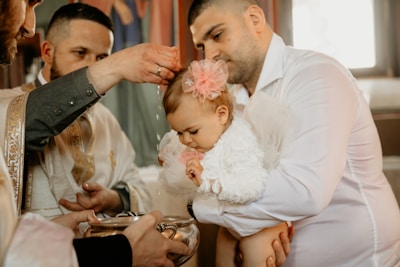 A baby dressed in white with a pink floral headband is being held by a man during a baptism ceremony. A priest is pouring water over the baby's head with a silver container. The setting appears religious, with ornate garments worn by the priest.