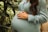 A person wearing a light blue dress is cradling their pregnant belly with both hands. Surrounding them are lush green plants.