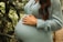 A person wearing a light blue dress is cradling their pregnant belly with both hands. Surrounding them are lush green plants.