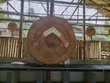 A traditional woven bamboo structure with a large circular bamboo hat displayed in the center, decorated with a red and white cloth. Smaller circular bamboo items hang in the background. The setting includes bamboo walls and a bench with some books laid out.