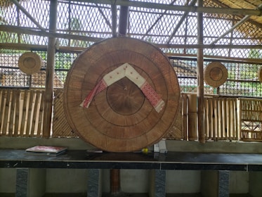 A traditional woven bamboo structure with a large circular bamboo hat displayed in the center, decorated with a red and white cloth. Smaller circular bamboo items hang in the background. The setting includes bamboo walls and a bench with some books laid out.