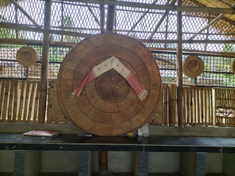A traditional woven bamboo structure with a large circular bamboo hat displayed in the center, decorated with a red and white cloth. Smaller circular bamboo items hang in the background. The setting includes bamboo walls and a bench with some books laid out.