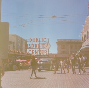 A bustling market scene with people walking along a cobblestone street. The iconic red sign of 'PUBLIC MARKET CENTER' is prominently displayed, with a vintage car parked in the background. The lively atmosphere is enhanced by the presence of vendors and vibrant red umbrellas.