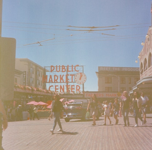 A bustling market scene with people walking along a cobblestone street. The iconic red sign of 'PUBLIC MARKET CENTER' is prominently displayed, with a vintage car parked in the background. The lively atmosphere is enhanced by the presence of vendors and vibrant red umbrellas.
