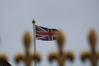A British flag is waving on a flagpole against a cloudy sky, with the top of a decorative gold-colored fence in the foreground.