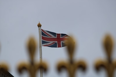 A British flag is waving on a flagpole against a cloudy sky, with the top of a decorative gold-colored fence in the foreground.
