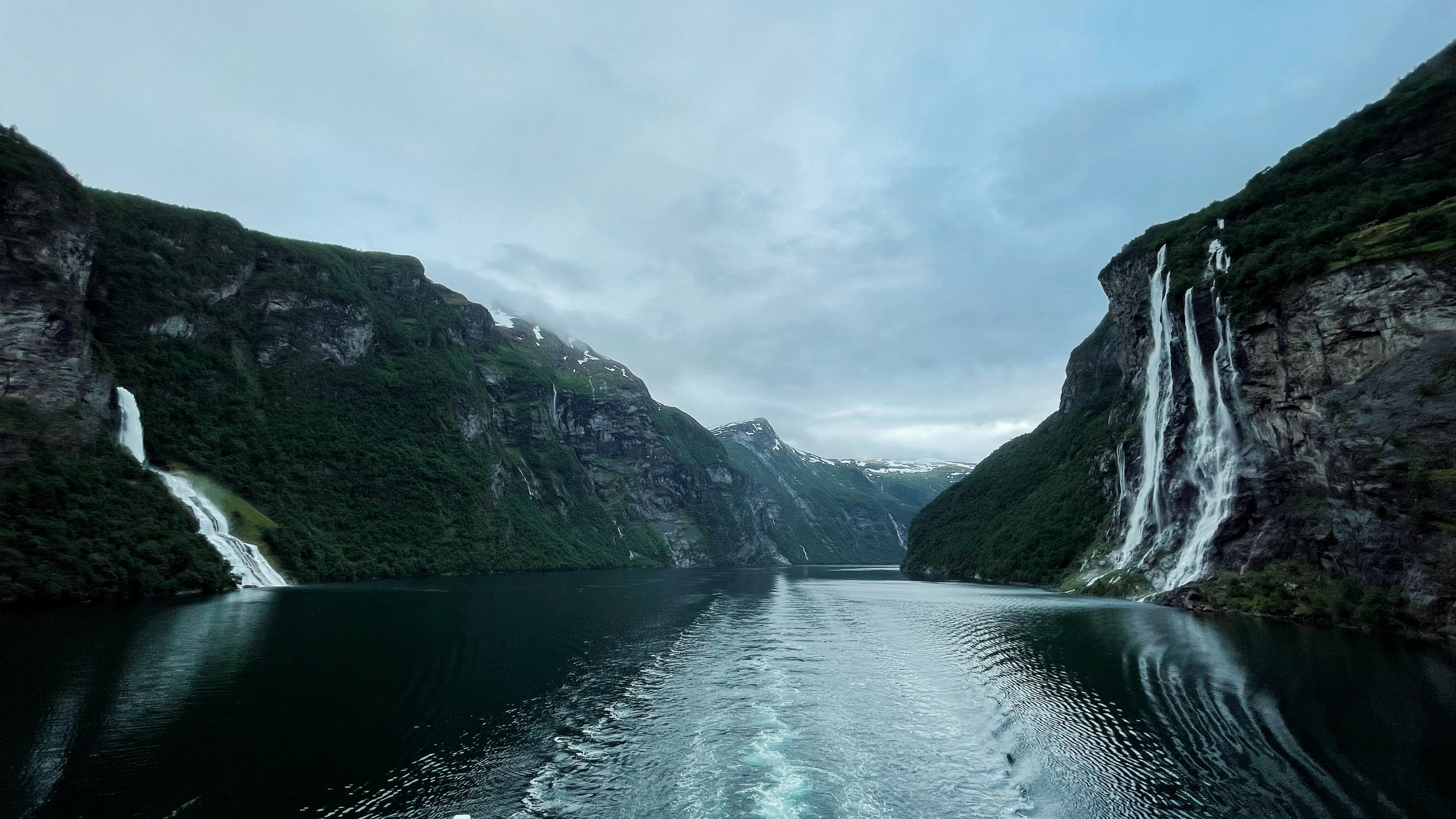 a body of water surrounded by mountains and a waterfall, Breathtaking view of Geirangerfjord and Seven Sisters waterfall made from a boat while sailing in silence.