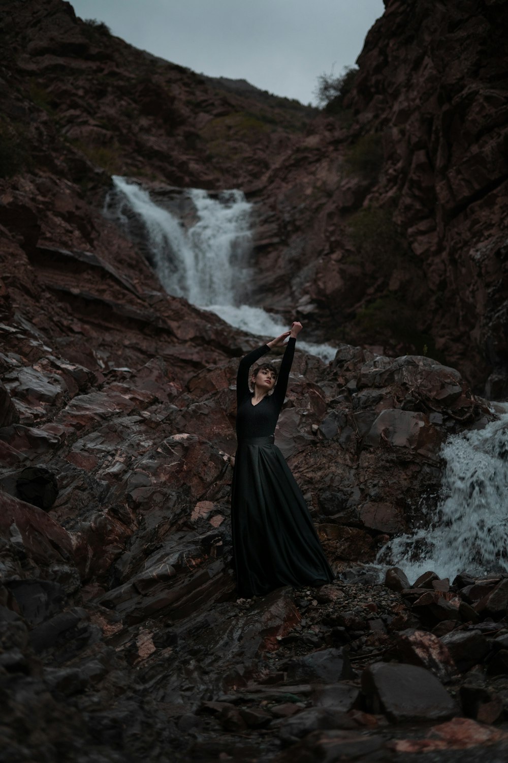 a woman in a long black dress standing on rocks in front of a waterfall