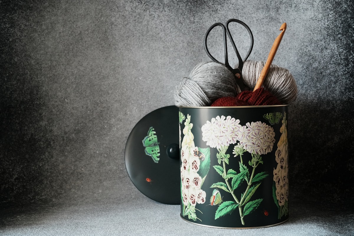 A decorative tin holding balls of yarn, a wooden crochet hook, and vintage scissors