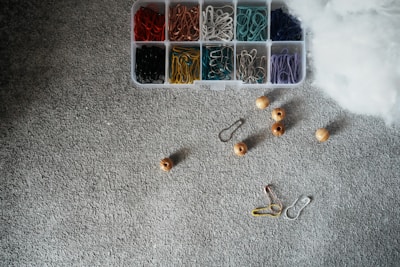A textured gray surface is scattered with a variety of crafting or organizational supplies. There is a plastic container divided into sections, each holding paper clips or hook-like fasteners in different colors including red, orange, green, yellow, silver, teal, purple, and black. Nearby, several wooden beads and metal clips are spread across the surface. A fluffy white material is partially visible on one side of the image.