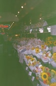 A market stall filled with numerous colorful flower bouquets, including sunflowers and other assorted blooms wrapped in paper. The setting appears to be indoors with dim lighting, and a neon sign hangs in the background. The bouquets are neatly arranged on tables creating a vibrant display.