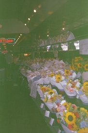 A market stall filled with numerous colorful flower bouquets, including sunflowers and other assorted blooms wrapped in paper. The setting appears to be indoors with dim lighting, and a neon sign hangs in the background. The bouquets are neatly arranged on tables creating a vibrant display.