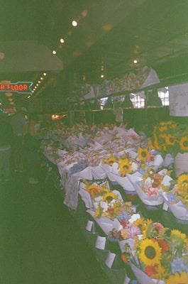 A market stall filled with numerous colorful flower bouquets, including sunflowers and other assorted blooms wrapped in paper. The setting appears to be indoors with dim lighting, and a neon sign hangs in the background. The bouquets are neatly arranged on tables creating a vibrant display.