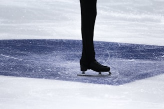 A person is standing on ice wearing black ice skates and dark trousers. The ice surface shows visible marks and scratches, indicating movement or skating. The area is well-lit, highlighting the texture of the ice.