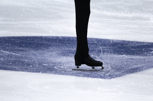 A person is standing on ice wearing black ice skates and dark trousers. The ice surface shows visible marks and scratches, indicating movement or skating. The area is well-lit, highlighting the texture of the ice.