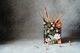 A fabric container with a floral design holds various crochet hooks and a pair of scissors. The container is surrounded by small wooden beads scattered on a textured gray surface.