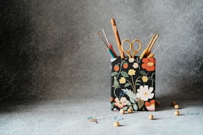 A fabric container with a floral design holds various crochet hooks and a pair of scissors. The container is surrounded by small wooden beads scattered on a textured gray surface.