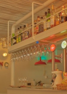 A bar counter featuring a hanging rack of wine glasses and a variety of liquor bottles arranged on a shelf above. The warm lighting creates an inviting atmosphere. In the background, red lampshades hang over additional glassware and plates, and a beverage dispenser with visible branding is on one side.