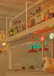 A bar counter featuring a hanging rack of wine glasses and a variety of liquor bottles arranged on a shelf above. The warm lighting creates an inviting atmosphere. In the background, red lampshades hang over additional glassware and plates, and a beverage dispenser with visible branding is on one side.