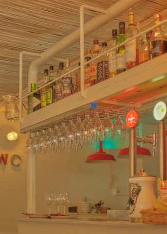 A bar counter featuring a hanging rack of wine glasses and a variety of liquor bottles arranged on a shelf above. The warm lighting creates an inviting atmosphere. In the background, red lampshades hang over additional glassware and plates, and a beverage dispenser with visible branding is on one side.