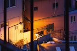 Urban setting featuring closely-packed buildings in twilight, viewed from behind bars. The scene includes warm artificial lighting illuminating building facades and creating vivid contrasts with shadows. Various architectural elements like windows and air conditioning units are visible, along with cables stretched across the walls.