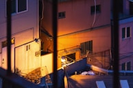 Urban setting featuring closely-packed buildings in twilight, viewed from behind bars. The scene includes warm artificial lighting illuminating building facades and creating vivid contrasts with shadows. Various architectural elements like windows and air conditioning units are visible, along with cables stretched across the walls.