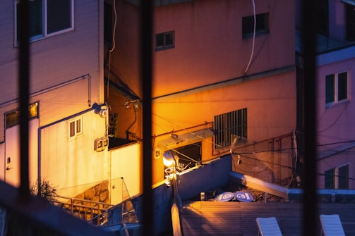 Urban setting featuring closely-packed buildings in twilight, viewed from behind bars. The scene includes warm artificial lighting illuminating building facades and creating vivid contrasts with shadows. Various architectural elements like windows and air conditioning units are visible, along with cables stretched across the walls.
