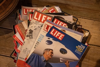 A stack of vintage Life magazines is arranged neatly in a black wire basket on a wooden floor. The covers showcase various themes with vibrant and bold imagery, featuring a mix of people and landscapes. Prominent titles and headlines accompany the images on the covers.