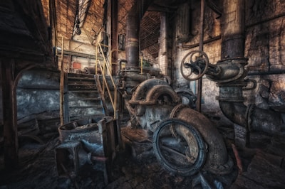 An abandoned industrial facility with large, rusted pipes and machinery. The structure appears aged and is covered in dust and grime. Dim lighting casts shadows across the surfaces, highlighting the intricate network of valves and metal parts.