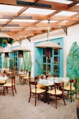 A charming outdoor dining area with wooden tables and chairs featuring yellow cushions. The setting includes teal-colored window frames and tropical leaf decorations on the walls. Overhead, woven light fixtures hang from wooden beams, complemented by greenery.