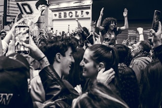 A lively crowd gathered in celebration, with people smiling and raising their hands. One individual stands atop the crowd, enthusiastically cheering. Camera phones capture the moment, and flags are partially visible. The setting appears to be on a street outside a store with a sign in the background.