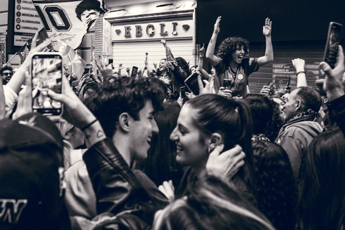 A lively crowd gathered in celebration, with people smiling and raising their hands. One individual stands atop the crowd, enthusiastically cheering. Camera phones capture the moment, and flags are partially visible. The setting appears to be on a street outside a store with a sign in the background.