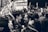 A lively crowd gathered in celebration, with people smiling and raising their hands. One individual stands atop the crowd, enthusiastically cheering. Camera phones capture the moment, and flags are partially visible. The setting appears to be on a street outside a store with a sign in the background.