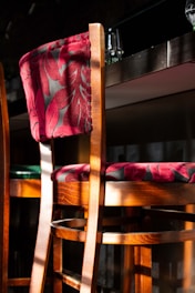 A wooden bar stool with a richly textured fabric backrest and seat, featuring a deep maroon color with a leaf pattern. The surface is bathed in warm sunlight, casting shadows and creating a cozy atmosphere. In the background, there are glass items on a countertop, hinting at a bar or a restaurant setting.