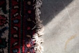 A textile rug with intricate patterns and vibrant colors lies partially on a cement floor. The rug features a mix of dark red, blue, black, and tan designs with a fringe of white threads at the edge. Sunlight casts shadows on the floor, creating contrasting textures.