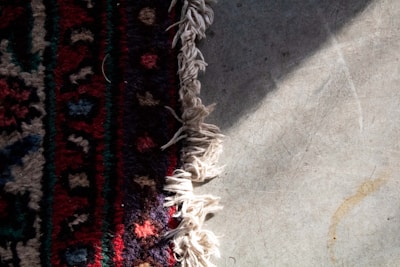 A textile rug with intricate patterns and vibrant colors lies partially on a cement floor. The rug features a mix of dark red, blue, black, and tan designs with a fringe of white threads at the edge. Sunlight casts shadows on the floor, creating contrasting textures.