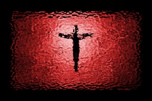 A textured depiction of a cross silhouette centered against a vibrant red and black background, resembling stained glass or a ripple effect.