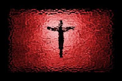 A textured depiction of a cross silhouette centered against a vibrant red and black background, resembling stained glass or a ripple effect.