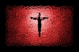 A textured depiction of a cross silhouette centered against a vibrant red and black background, resembling stained glass or a ripple effect.