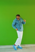 A person wearing a denim shirt, white pants, and blue sneakers stands against a vibrant green background. The person has braided hair and is wearing sunglasses, striking a confident pose.