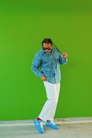 A person wearing a denim shirt, white pants, and blue sneakers stands against a vibrant green background. The person has braided hair and is wearing sunglasses, striking a confident pose.