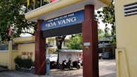 Entrance gate of a school labeled 'Hòa Vang' with large pillars and a sign displaying the school's name. Trees provide shade over the entrance, and there are motorcycles parked in the area. A yellow wall runs alongside, featuring some text. In the background, parts of buildings and a car are visible, indicating an urban setting.