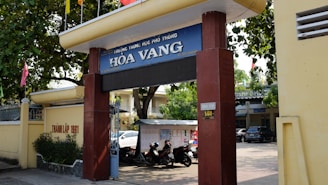 Entrance gate of a school labeled 'Hòa Vang' with large pillars and a sign displaying the school's name. Trees provide shade over the entrance, and there are motorcycles parked in the area. A yellow wall runs alongside, featuring some text. In the background, parts of buildings and a car are visible, indicating an urban setting.