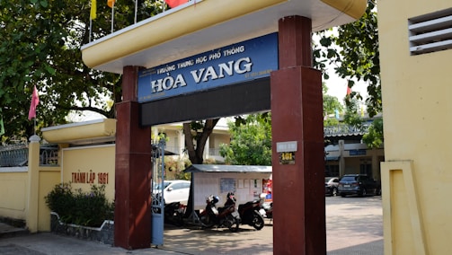 Entrance gate of a school labeled 'Hòa Vang' with large pillars and a sign displaying the school's name. Trees provide shade over the entrance, and there are motorcycles parked in the area. A yellow wall runs alongside, featuring some text. In the background, parts of buildings and a car are visible, indicating an urban setting.